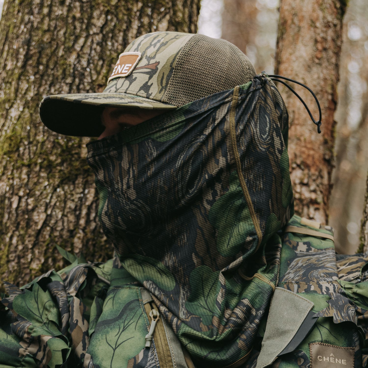 Person in full camouflage gear with a mask, sitting against a tree in a forest.