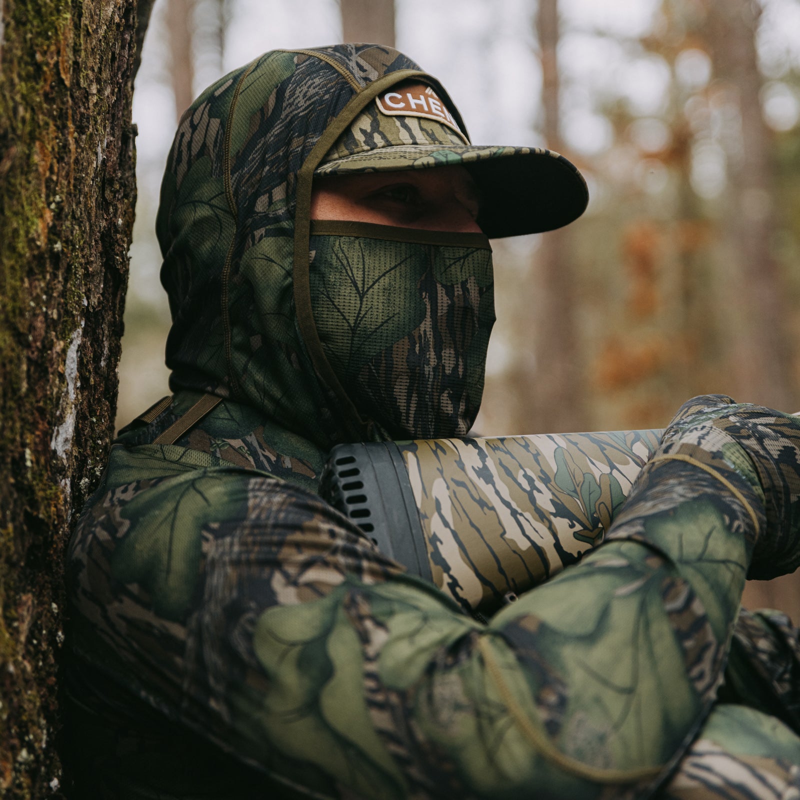 Person in full camouflage gear sitting against a tree in a forest