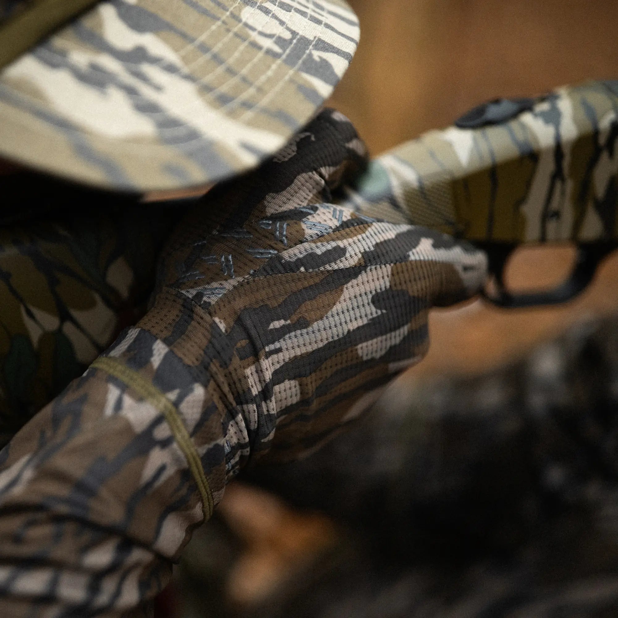 Camouflage-patterned glove holding a rifle, with a blurred background #mossy_oak_original_bottomland